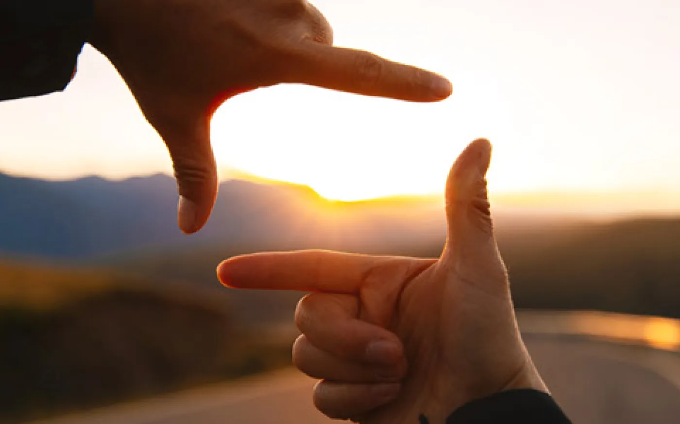 Two hands forming a rectangular frame around a sunset over a winding road and mountains.