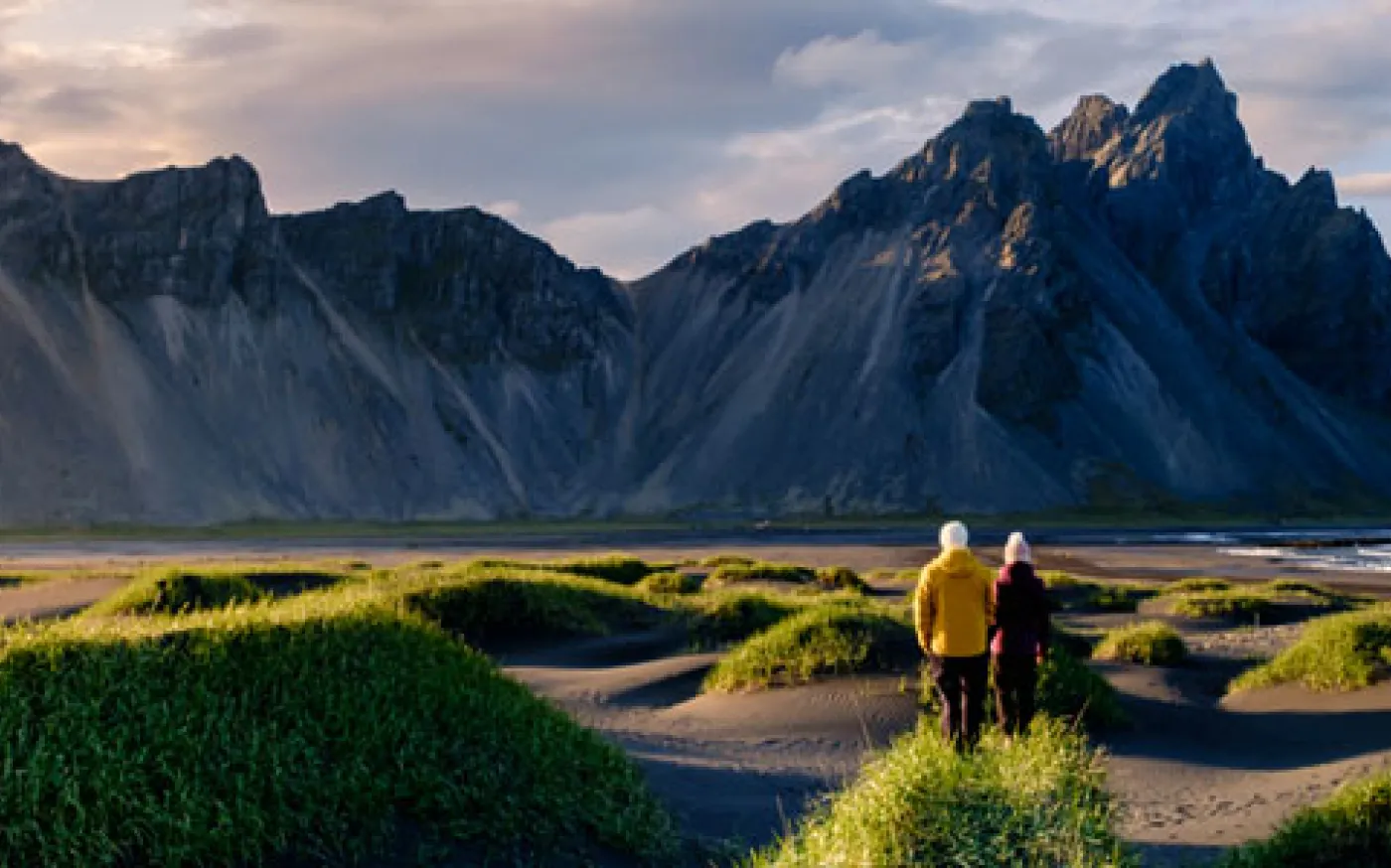 Two people walking through a grassy coastal landscape toward dramatic rocky mountains.