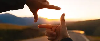 Two hands forming a rectangular frame around a sunset over a winding road and mountains.